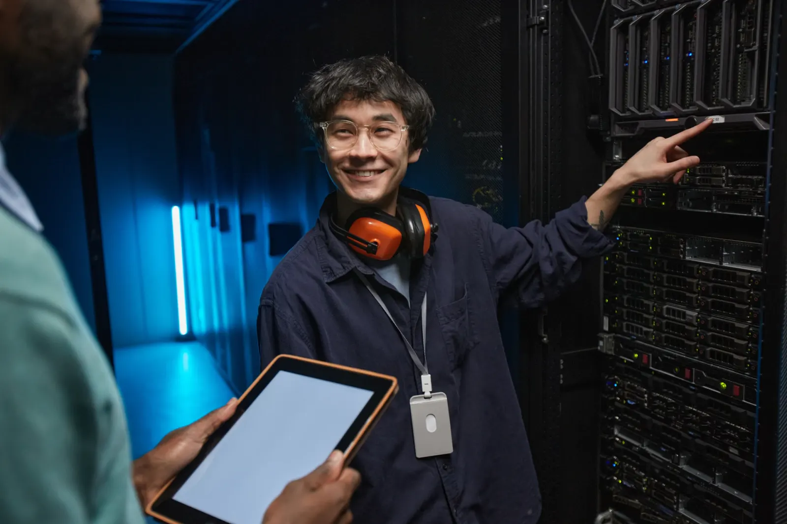 Two engineers in a data centre, one pointing out a component on a server rack while the other holds a tablet.