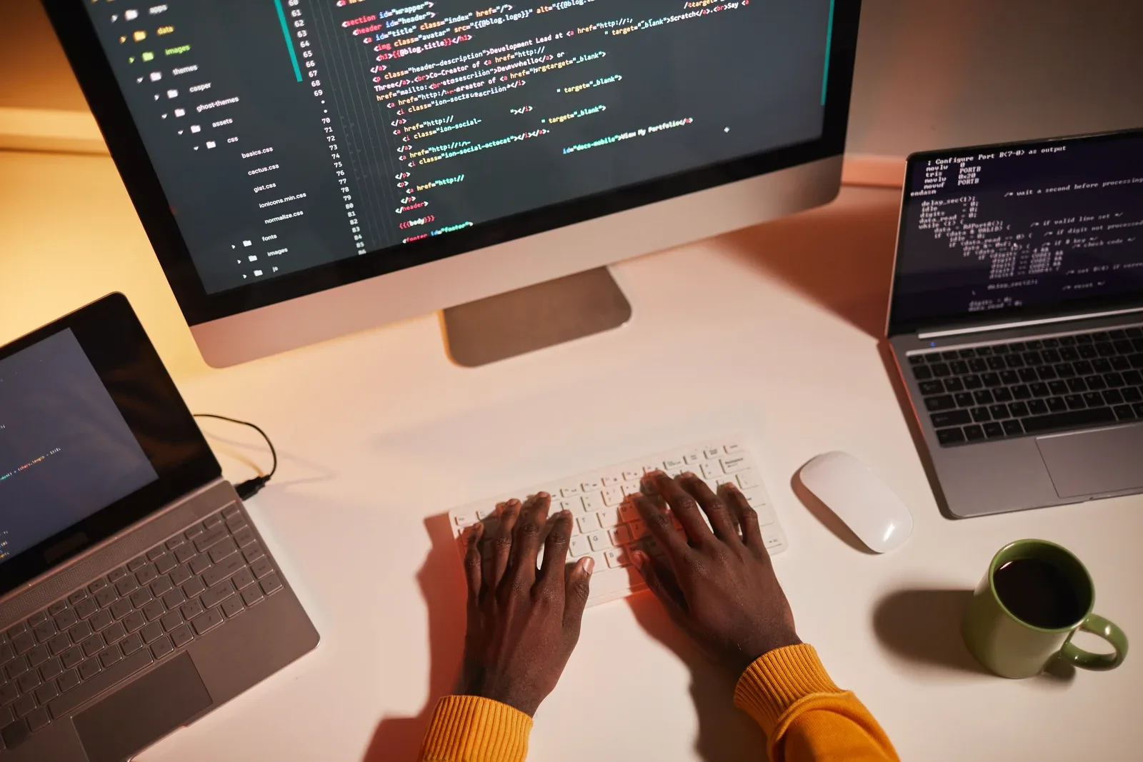Hands typing at a warm-lit desk between a monitor, laptop and tablet, all showing code.