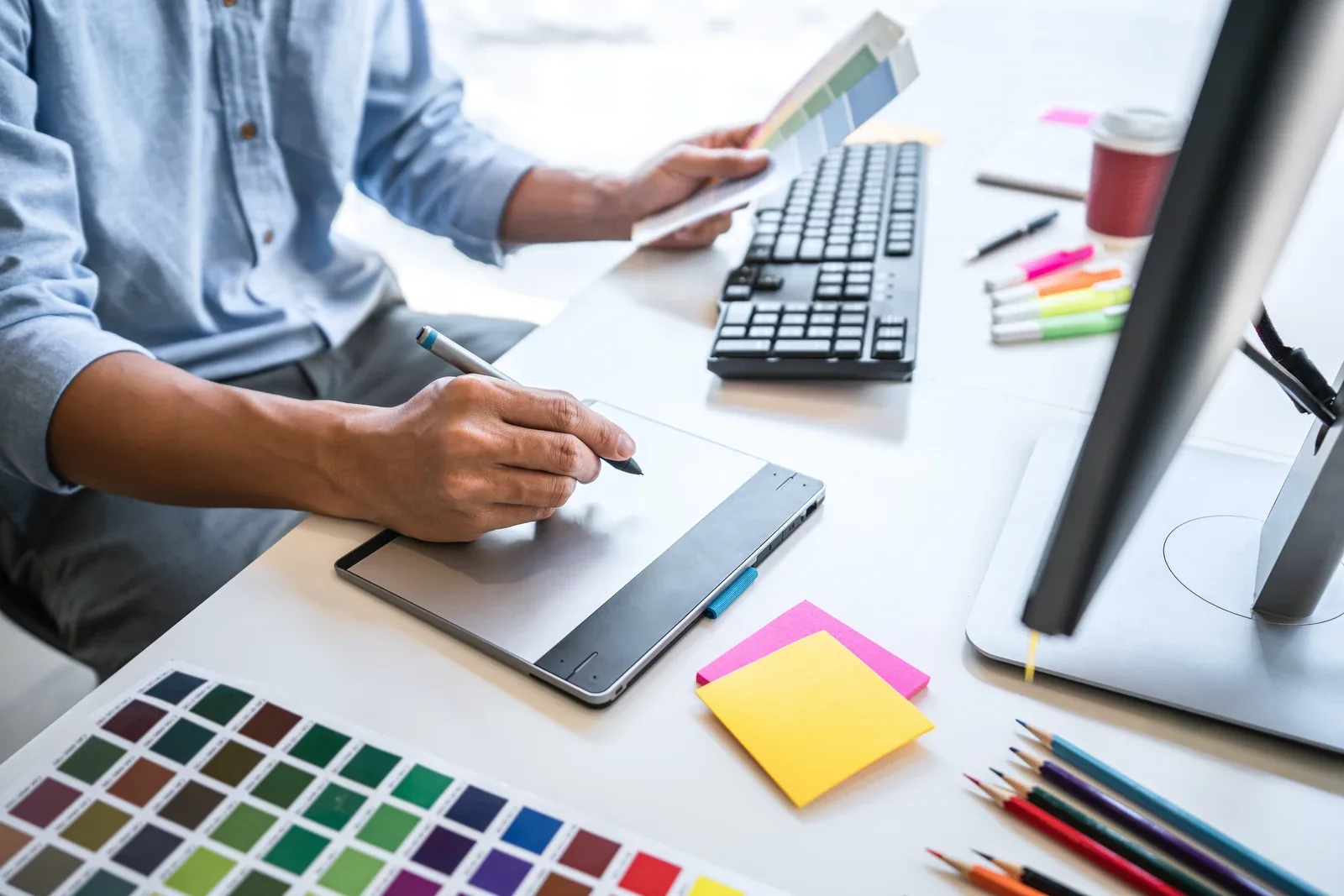 A designer sketching on a graphics tablet beside printed colour swatches and pencils.