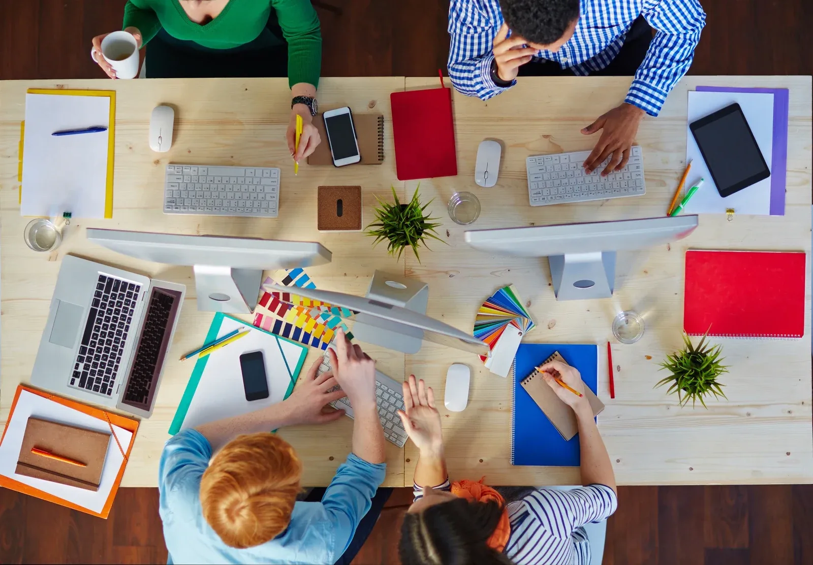 Overhead view of a creative team collaborating at a shared desk covered in computers, colour swatches, notebooks and phones.