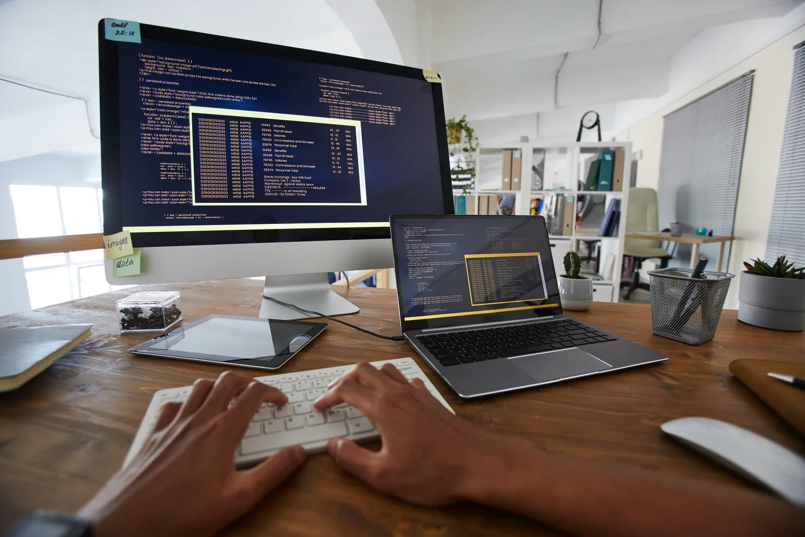 A developer's workspace on a bright desk, with code across a desktop monitor and two laptop screens.
